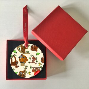 May include: A round, cream-colored ornament with a red ribbon. The ornament features a repeating pattern of orangutans in various poses, including swinging from branches and holding bananas. The text "Selina Jayne" is printed in red on the ornament.