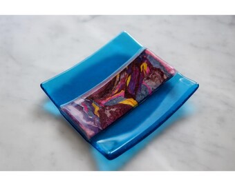 May include: A square, turquoise-blue glass dish with a rectangular, multi-colored accent piece. The accent piece features swirls of purple, yellow, and maroon. The dish is likely used for decorative purposes.