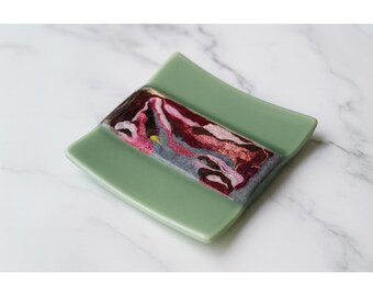 May include: A square, sage green glass dish with a rectangular, abstract design in the center. The design features swirls of burgundy, pink, gray, and yellow. The dish has slightly curved edges and a smooth surface.