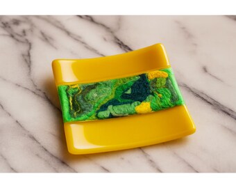 May include: A square, yellow soap dish with a central band of green and yellow marbled design. The dish has a glossy finish and is set against a marble background. The soap dish is a decorative item.