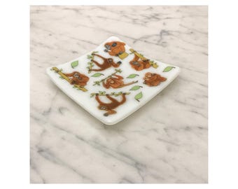 May include: Square white dish with a diamond shape, featuring cartoon orangutans hanging from branches with green leaves. The orangutans are in various poses, and the dish is set on a marble surface.