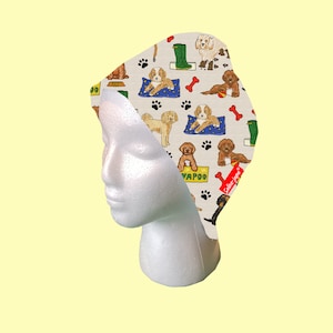以下が含まれることがあります： 白地に茶色と白の犬、骨、犬に関連する言葉の模様がプリントされた白い布製のシュシュ。シュシュは白いマネキンの頭に置かれている。