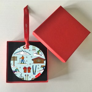 以下が含まれることがあります： 冬スポーツをテーマにした、丸い青と白のクリスマスオーナメント。オーナメントには、スキーヤー、スキーロッジ、スキー用品が描かれています。オーナメントは、黒い内装の赤い箱に入っています。