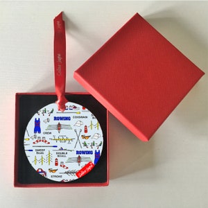 May include: A white round ornament with a blue and red rowing themed design. The ornament features illustrations of rowing equipment, a boat, and the words "ROWING" and "Selina-Jame".