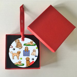 May include: A round white ceramic ornament with a red ribbon. The ornament features a pattern of brown and white doodle dogs doing yoga poses. The text "A cute cockapoo lives here" is on a blue sign.