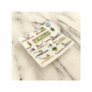 May include: A white, square trinket dish with a Pilates theme. The dish features illustrations of Pilates poses, the word "PILATES", and the words "Flow", "Control", and "Flexibility". The dish is decorated with green, yellow, and blue accents.