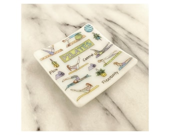 May include: A white, square trinket dish with a Pilates theme. The dish features illustrations of Pilates poses, the word "PILATES", and the words "Flow", "Control", and "Flexibility". The dish is decorated with green, yellow, and blue accents.