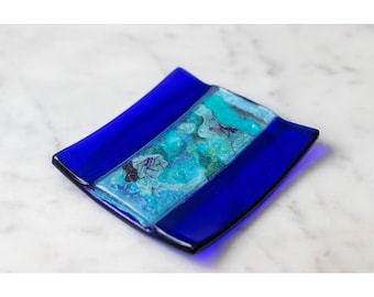 May include: A square, cobalt blue glass dish with a central rectangular section featuring turquoise and teal accents. The dish has a wavy edge and is likely handmade, showcasing a unique artistic design.