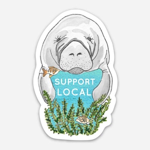 May include: A sticker featuring a watercolor illustration of a manatee with the text "SUPPORT LOCAL" in blue. The manatee is gray and white, with a fish in its mouth and aquatic plants at the bottom.