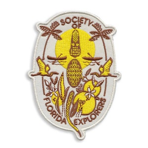 May include: A white oval embroidered patch with the text "Society of Florida Explorers" in brown lettering. The patch features a yellow sun with a brown alligator in the center, surrounded by palm trees, birds, and flowers.