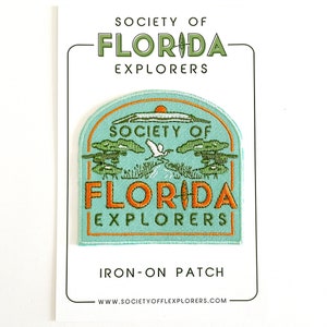 May include: A turquoise blue iron-on patch with an orange border. The patch features a white bird flying over a green tree and the text "Society of Florida Explorers".