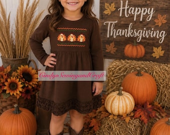 May include: A long-sleeved brown dress featuring a smocked chest panel with a repeating turkey pattern, a ruffled hem, and a dark brown hair bow. The dress includes a small tag with the text Cindy's SewingandCraft.