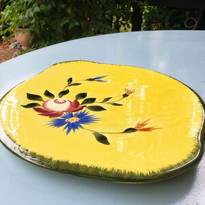 May include: A yellow ceramic serving platter with a hand-painted floral design. The flowers include a red rose, blue and yellow flowers, and green leaves. The platter has a dark green rim.