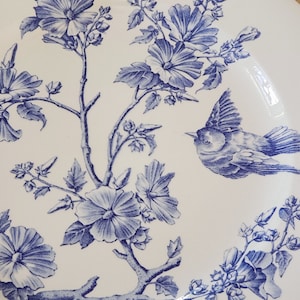 Puede incluir: Un plato de cerámica blanco con un diseño floral azul y un pájaro azul. Las flores son estilizadas y tienen un aspecto delicado y vintage.