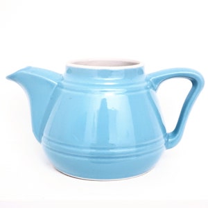 Peut inclure: Une théière en céramique bleu clair avec un corps arrondi et un bec large. La théière a un intérieur blanc et un bord blanc autour du haut.