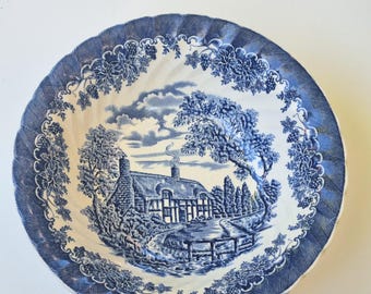 May include: Blue and white ceramic plate with a detailed landscape scene. The plate features a rural cottage, trees, and a river, framed by a decorative border of vines and a scalloped edge. A classic, vintage design.