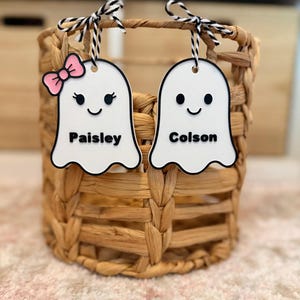 May include: Two white ghost-shaped ornaments with black outlines and black and white twine hangers. One ghost features a pink bow, eyelashes, and the name Paisley. The other ghost features the name Colson. Both have black eyes and smiles.