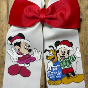 May include: A festive hair bow with a red bow and white ribbons. The ribbons feature cartoon characters: Minnie Mouse in a red dress and Santa hat, and Mickey Mouse and Pluto in festive sweaters.