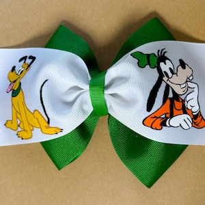 May include: A handmade hair bow featuring Pluto and Goofy. The bow has a white center with the characters printed on it, and is layered over a green ribbon base. The bow is tied with a green center knot.