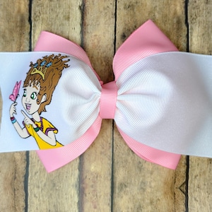 May include: A large, layered hair bow featuring a cartoon illustration of a girl with brown hair and a yellow dress. The bow has a white center, pink accents, and a pink butterfly.
