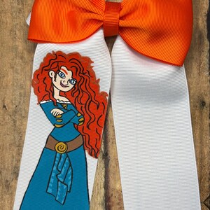 May include: A large hair bow featuring a cartoon illustration of a character with red curly hair, wearing a blue dress. The bow has an orange top section and white ribbons. The bow is made of a grosgrain ribbon.