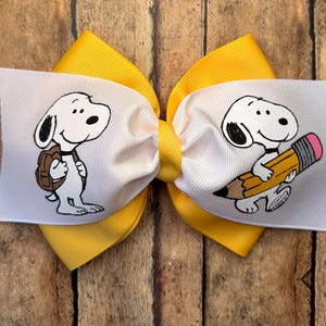 May include: A hair bow featuring a bright yellow ribbon base layered with white ribbon panels. The white panels display a cartoon dog character, one side showing the dog with a brown backpack and the other side showing the dog holding a yellow pencil.