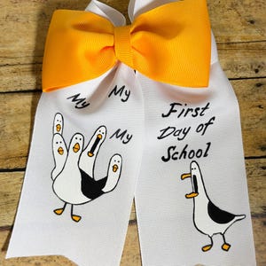 Peut inclure: Un nœud de cheveux en ruban blanc avec un nœud orange sur le dessus. Les queues de ruban présentent des mouettes de dessin animé et le texte "My First Day of School."