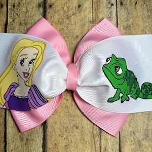 May include: A large, decorative hair bow featuring a pink and white layered design. The bow has a cartoon image of a blonde woman on one side and a green chameleon on the other. The bow is made of fabric.
