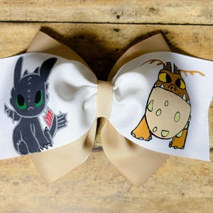 May include: A large hair bow featuring a white and tan color scheme. The bow has two cartoon dragon characters printed on the white ribbon. The bow is made of layered ribbons and is suitable for children.