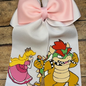 May include: A large white hair bow with a pink center bow and white streamers. The streamers feature cartoon illustrations of Princess Peach and Bowser from the Super Mario video game series.