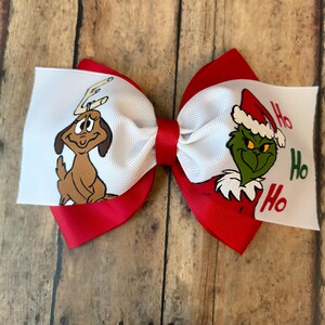 May include: A festive red and white hair bow featuring cartoon characters. One side displays a brown dog with antlers, and the other side shows a green character in a Santa hat with the words "Ho Ho Ho".