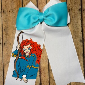 May include: A large white hair bow with a turquoise center and tails. One tail features a cartoon illustration of a character with red hair, wearing a blue dress and holding a bow and arrow. The bow is made of grosgrain ribbon.