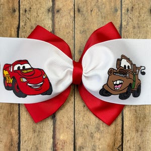 May include: A red and white hair bow featuring Lightning McQueen and Mater from the Cars movie. The bow has a red base with white ribbon on top, each displaying a character. The bow is set against a wooden background.
