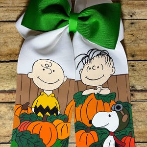 May include: A large hair bow featuring a green and white design. The bow has two white streamers with cartoon characters and pumpkins. Charlie Brown and Linus are featured, along with Snoopy. The bow is perfect for fall.