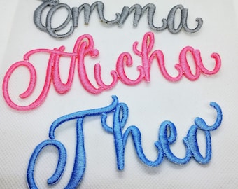 May include: Three embroidered names, Emma in gray, Micha in pink, and Theo in blue, on a white background.