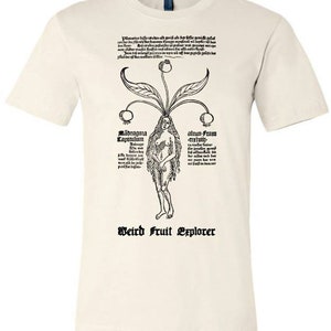 以下が含まれることがあります： 白いTシャツに、頭から果物が生えている女性の白黒のイラストが描かれています。女性の両側に「Mandragora Capitulum」と「Alrun-Frau -cretivu-」の文字があります。Tシャツの下部には「Weird Fruit Explorer」の文字があります。