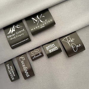 Peut inclure: Une collection d'étiquettes en tissu noir avec du texte blanc. Les étiquettes présentent diverses phrases, notamment "Rêver Grand", "Cocolinna", "Pequeños Encantos", "Take One", "petites beautés" et "Made with Love by Amanda".