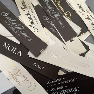 May include: A close-up of a pile of white and black fabric labels with gold lettering. The labels are for various businesses, including "Scarlet Thomas Interiors", "Amanda Torres", "Collin Anderson Collections", and "NOLA".