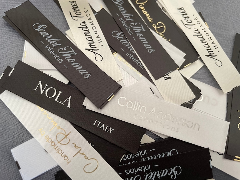 May include: A close-up of a pile of white and black fabric labels with gold lettering. The labels are for various businesses, including "Scarlet Thomas Interiors", "Amanda Torres", "Collin Anderson Collections", and "NOLA".
