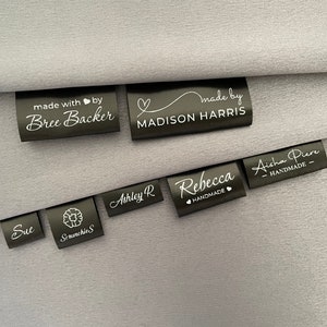 Può includere: Una collezione di etichette in raso nero con testo bianco. Le etichette presentano nomi e frasi come "made with love by Bree Backer", "made by MADISON HARRIS", "Sue", "Ashley R", "Srunchies", "Rebecca HANDMADE" e "Aisha Piere HANDMADE".