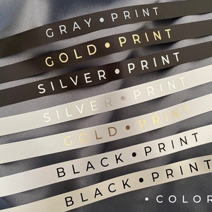 May include: A close-up of seven ribbon samples with different colors and text. The colors are gray, gold, silver, and black. The text on each ribbon reads "GRAY PRINT", "GOLD PRINT", "SILVER PRINT", "SILVER PRINT", "GOLD PRINT", "BLACK PRINT", and "BLACK PRINT". The last ribbon also has the text "&bull; COLORS." printed on it.