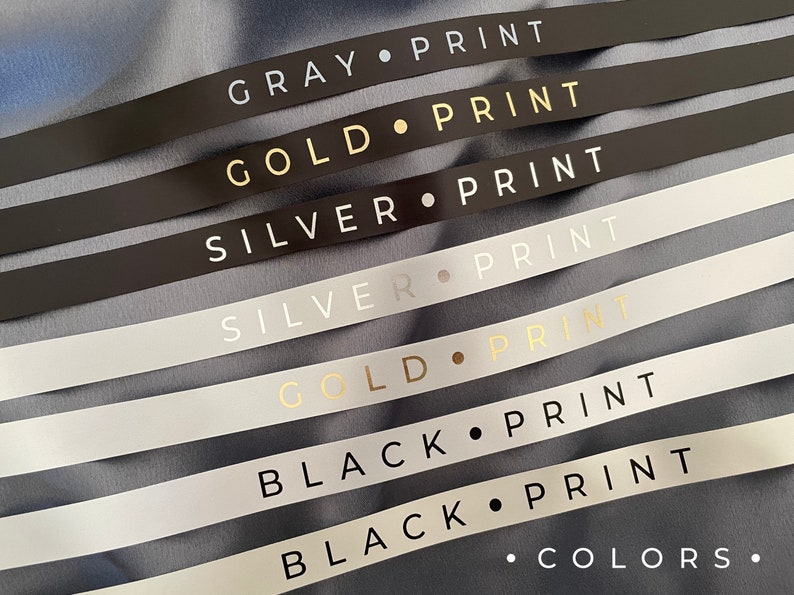 May include: A close-up of seven ribbon samples with different colors and text. The colors are gray, gold, silver, and black. The text on each ribbon reads "GRAY PRINT", "GOLD PRINT", "SILVER PRINT", "SILVER PRINT", "GOLD PRINT", "BLACK PRINT", and "BLACK PRINT". The last ribbon also has the text "&bull; COLORS." printed on it.