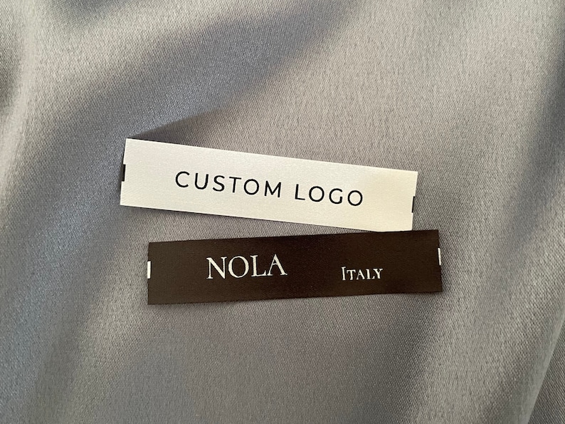 May include: Two white fabric labels with black text. The top label says "CUSTOM LOGO". The bottom label says "NOLA ITALY".