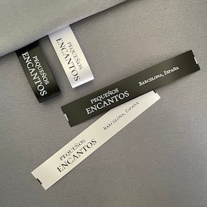 May include: Close-up of several fabric labels with the text "PEQUEÑOS ENCANTOS" in a serif font. Some labels are black with white text, others are white with black text. One label reads "BARCELONA, ESPAÑA".