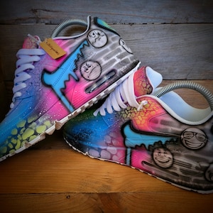 May include: A pair of custom-painted Nike Air Max 90 trainers with a colourful, abstract design. The shoes feature a blue, pink and green colour scheme with black accents. The design includes a variety of smiley faces and other graffiti-style elements.