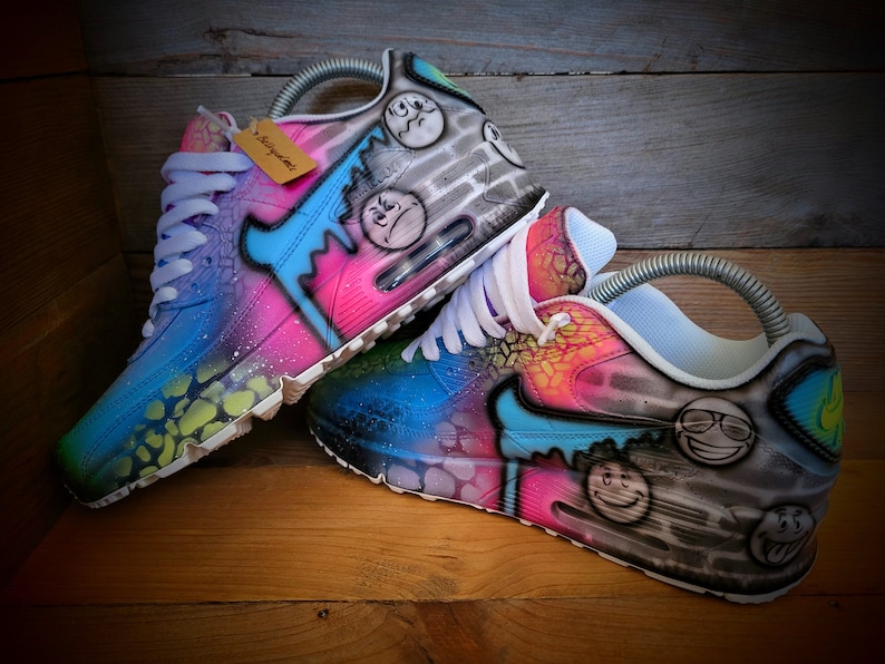 May include: A pair of custom-painted Nike Air Max 90 trainers with a colourful, abstract design. The shoes feature a blue, pink and green colour scheme with black accents. The design includes a variety of smiley faces and other graffiti-style elements.