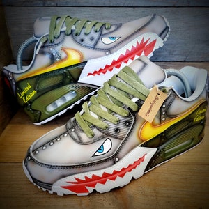 May include: A pair of custom-painted Nike Air Max 90 sneakers with a camouflage pattern, yellow accents, and a shark tooth design. The sneakers have a green and white color scheme and say "Limited Edition" on the side.