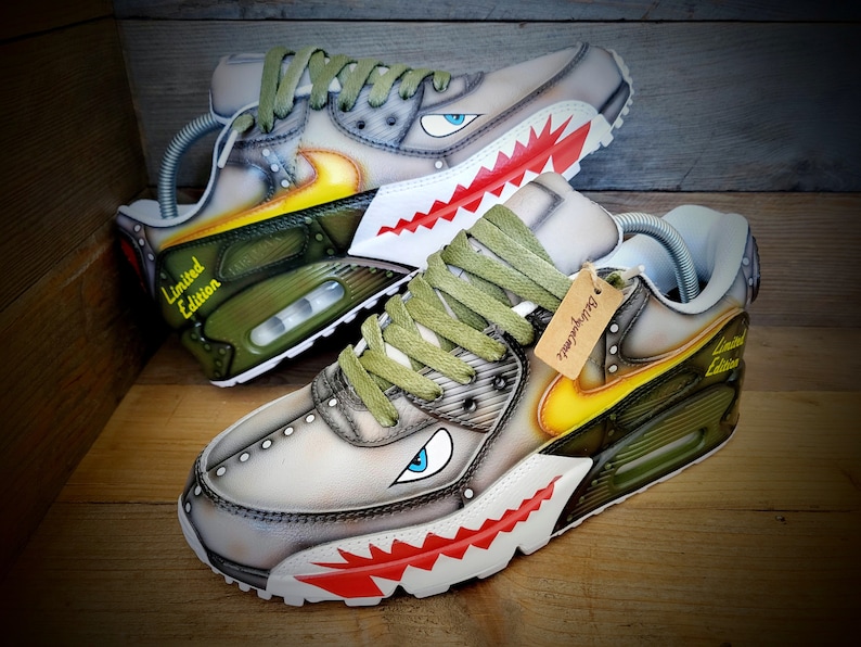 May include: A pair of custom-painted Nike Air Max 90 sneakers with a camouflage pattern, yellow accents, and a shark tooth design. The sneakers have a green and white color scheme and say "Limited Edition" on the side.