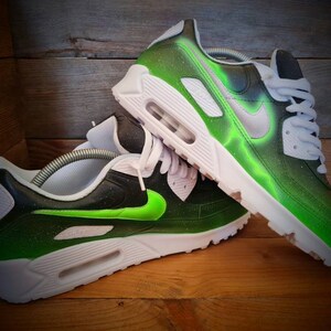 May include: A pair of custom-painted Nike Air Max 90 sneakers. The sneakers are black with green accents and a white swoosh logo. The green accents are a gradient, fading from a bright green to a darker green.