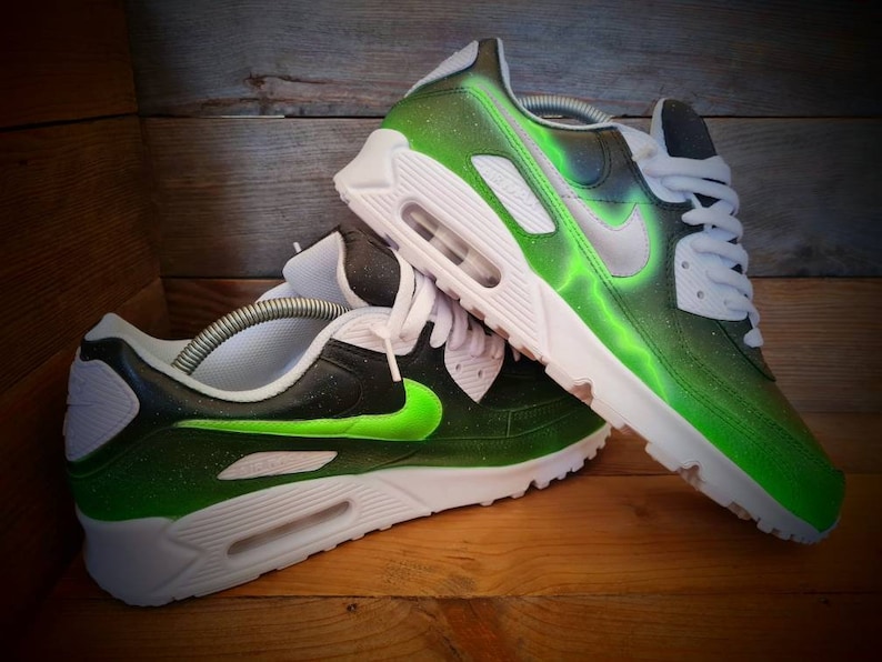 May include: A pair of custom-painted Nike Air Max 90 sneakers. The sneakers are black with green accents and a white swoosh logo. The green accents are a gradient, fading from a bright green to a darker green.